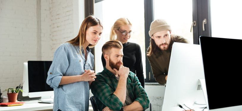 Photo of young serious colleagues in office talking with each other using computer. Looking aside.